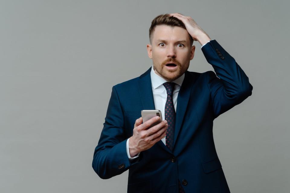 Shocked investor with mouth open, hand on head and smartphone in other hand.